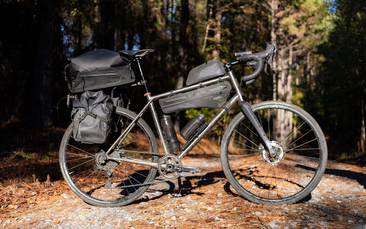A gray gravel bicycle loaded with black pannier bags, a top box, and a frame bag, parked on a forest path with pine trees in the background.