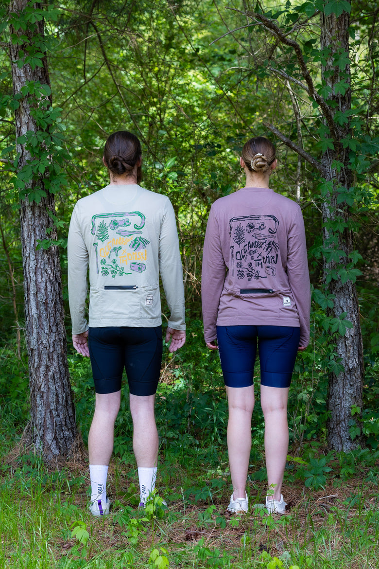 Two people standing with their backs to the camera in a wooded outdoor setting, wearing matching long-sleeve athletic shirts with large graphic prints on the back and dark bike shorts.