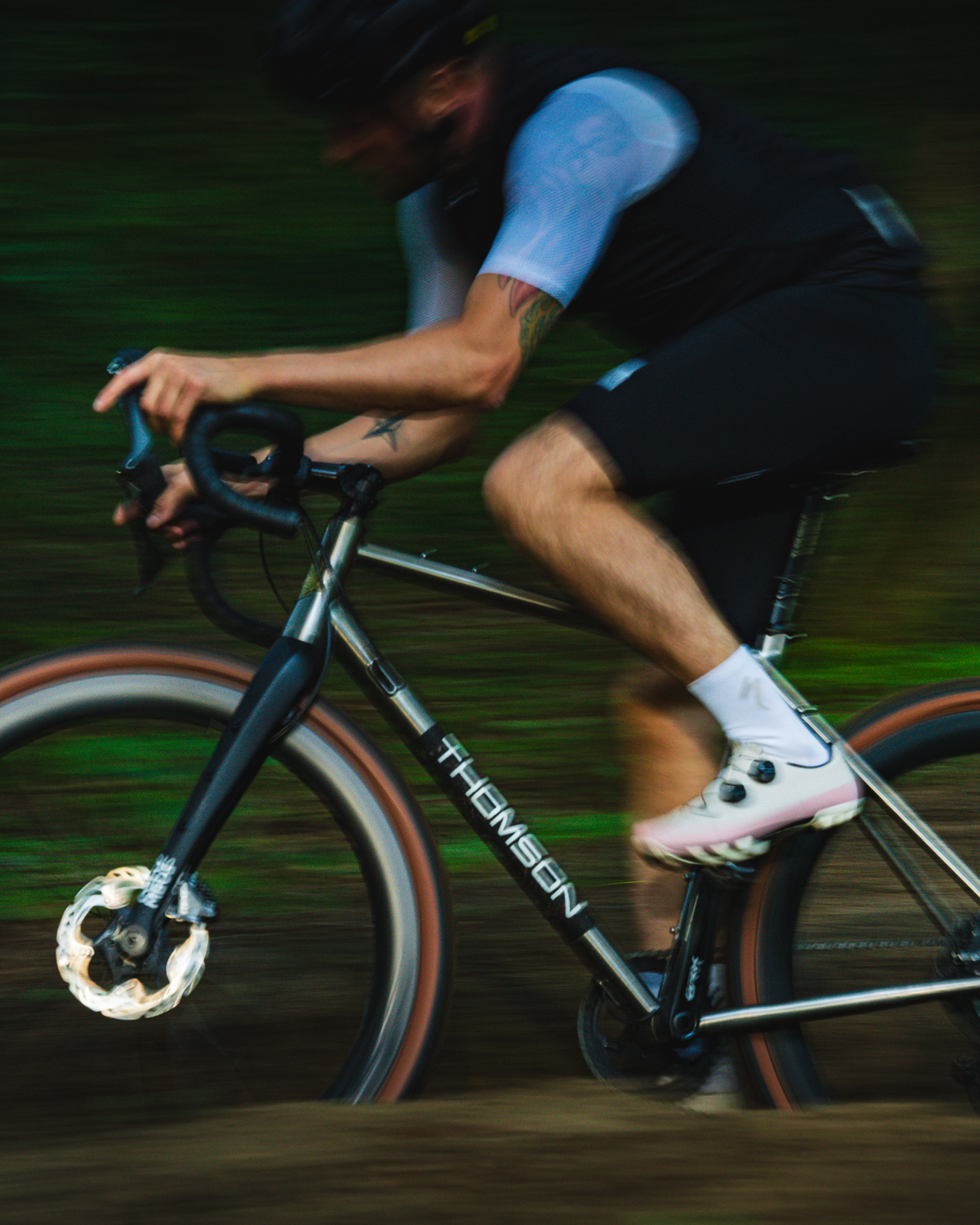A cyclist in black and white cycling gear rides a black Thomsen road/gravel bike on a dirt path, captured with a motion-blur effect that emphasizes speed.