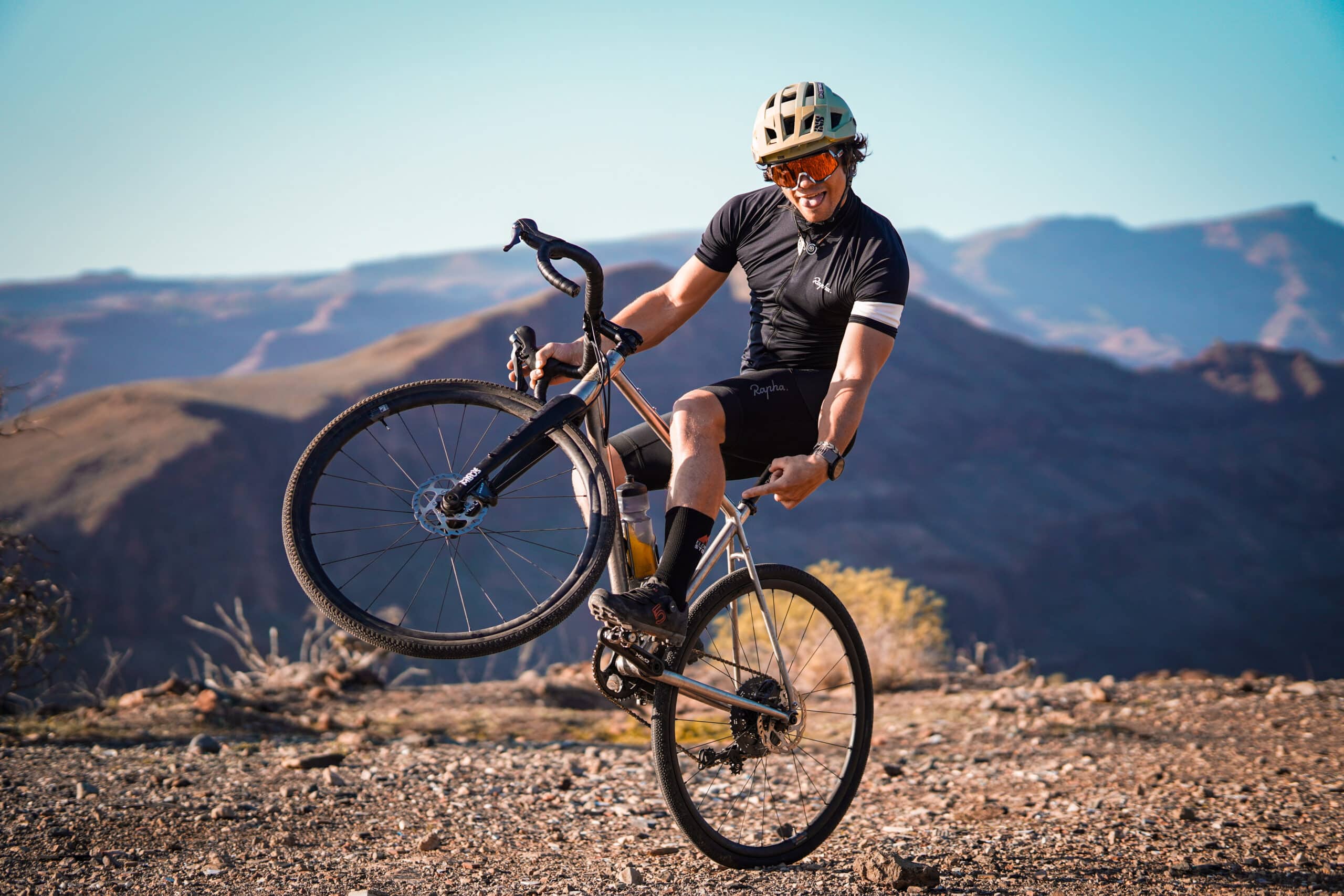 A cyclist wearing a helmet and sunglasses performs a wheelie on a gravel bike, smiling with a mountainous landscape in the background under a clear blue sky.