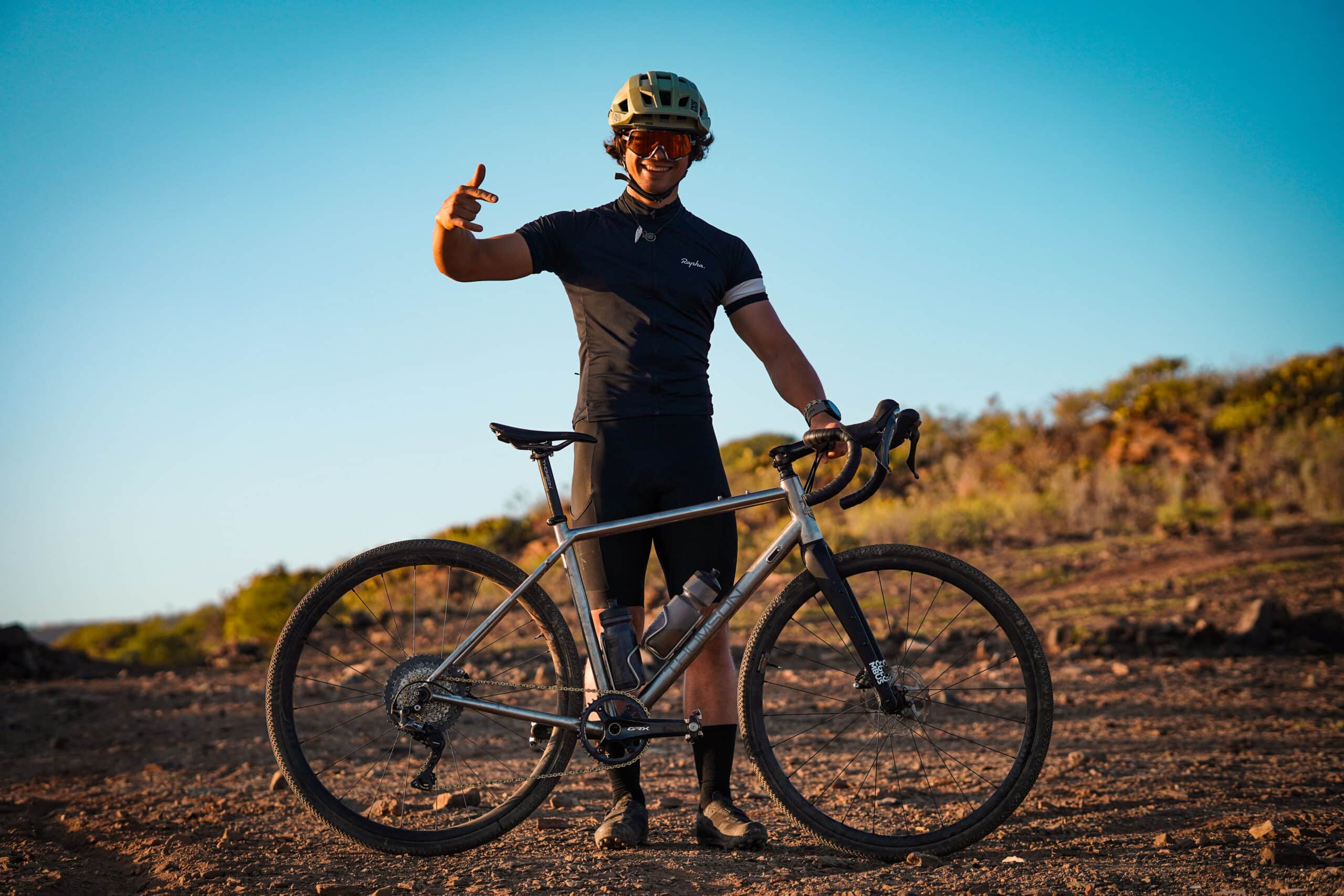  cyclist wearing a helmet and sunglasses stands on a dirt path with a gravel bike, smiling and giving a thumbs-up gesture under a clear blue sky.