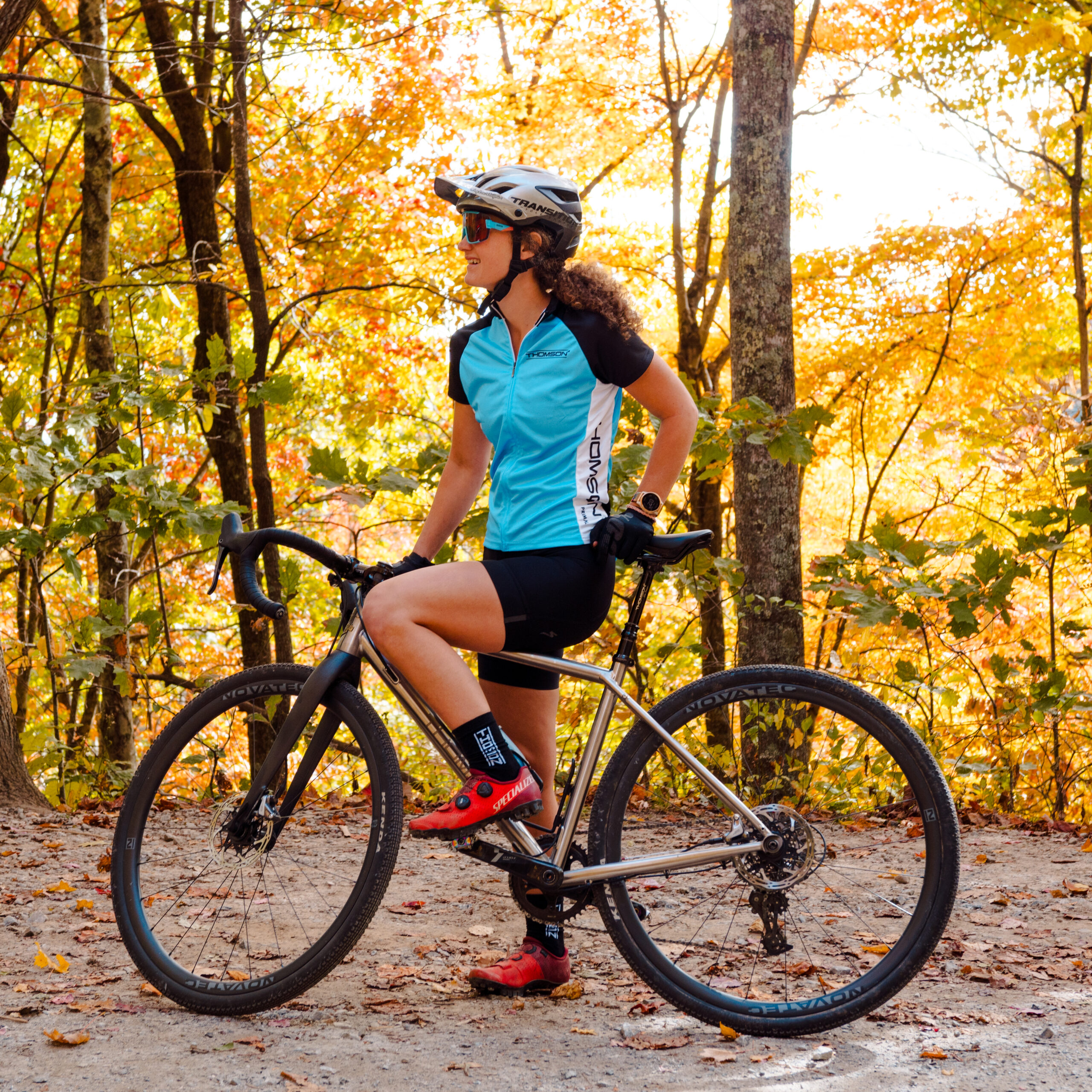 A cyclist wearing a blue and black jersey, helmet, and red cycling shoes stands with their bike on a dirt path in a forest with vibrant autumn foliage.